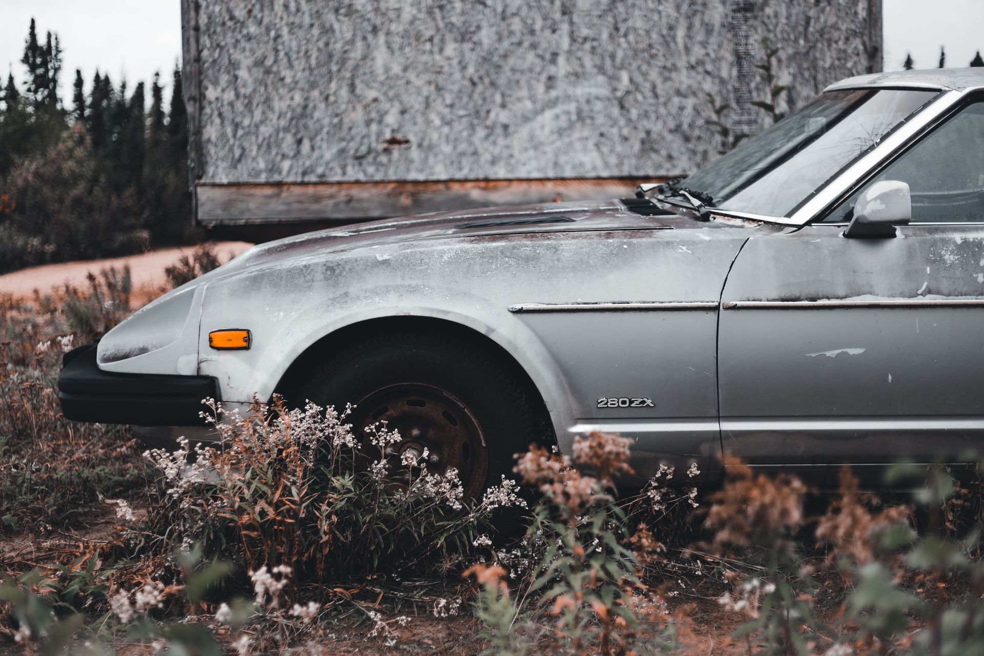 A classic Nissan 280ZX car parked outdoors, surrounded by overgrown weeds, showcasing a rustic and nostalgic vibe.